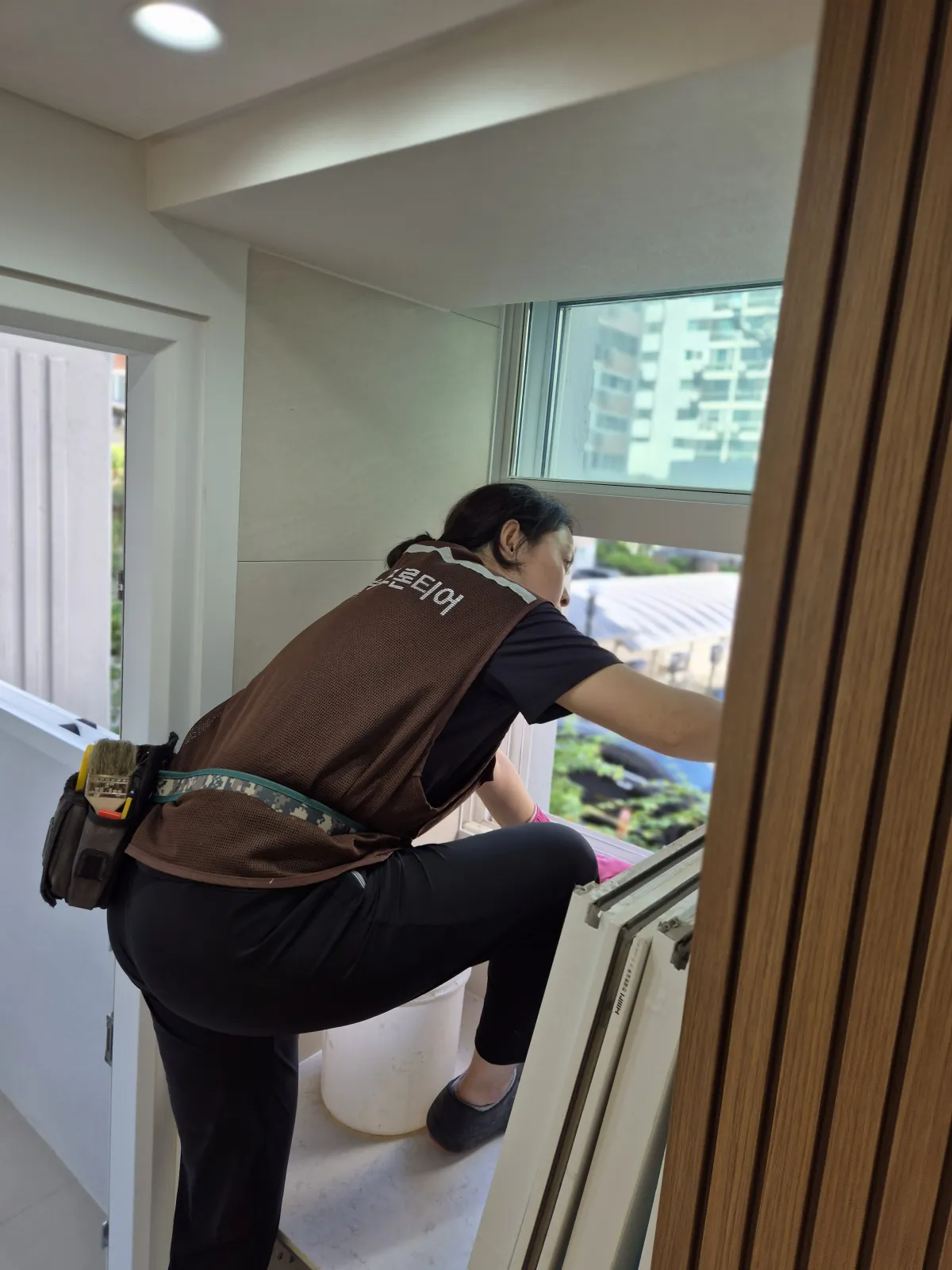 협력업체 조끼를 입은 작업자가 창을 넘어 새시 레일을 손으로 훑는 모습